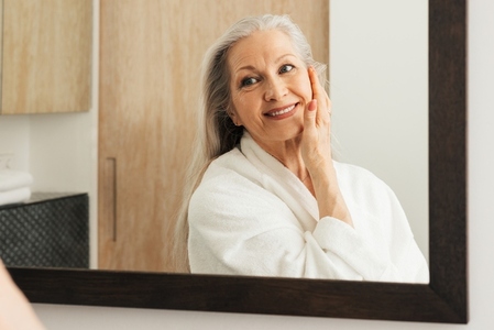 Senior woman with long grey hair touching her face with hand and looking at mirror
