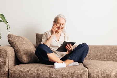 Smiling senior woman in eyeglasses sitting on a couch with a digital tablet