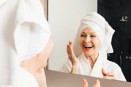 Laughing aged woman with a wrapped towel on her head applying cream on her face in bathroom