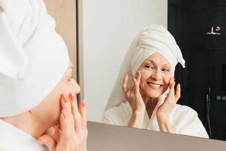 Smiling aged woman with a towel on a head applying facial cream on her cheeks in bathroom
