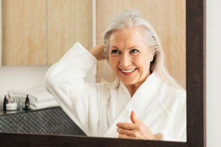 Cheerful aged woman in bathrobe adjusting her grey hair in front of a mirror