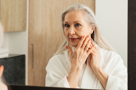 Senior woman with long grey hair touching her cheek with fingers in bathroom