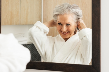 Smiling aged woman making funny hairstyle while looking at a mirror in bathroom  Senior female enjoying morning routine in the bathroom