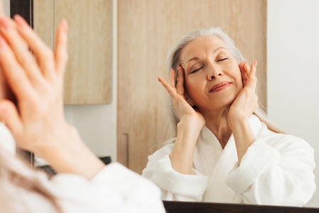 Aged woman with closed eyes touching her face with her fingers  Female in bathrobe doing face massage in bathroom