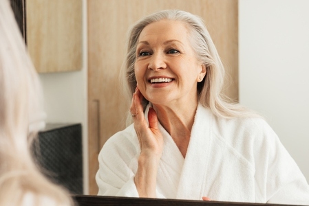 Happy aged female touching her face in front of a mirror  Woman in bathrobe admires her reflection