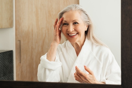 Cheerful aged woman touching her face in front of a bathroom mirror