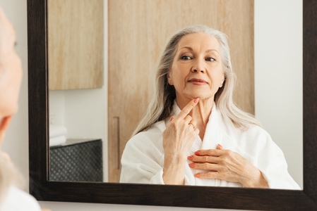 Portrait of a senior woman with grey hair touching her chin with a finger in front of a mirror