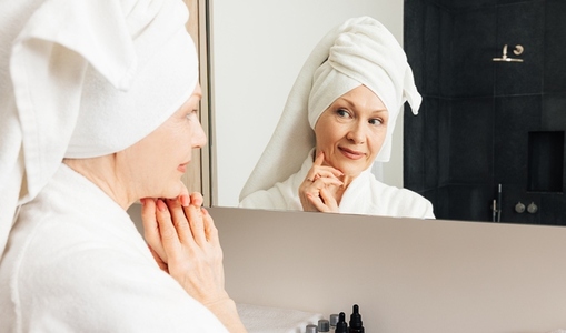 Aged female with a wrapped towel on a head looking at the bathroom mirror