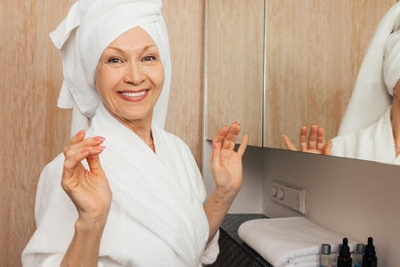 Smiling aged woman with a towel on her head looking at camera in a bathroom  Cheerful senior female in a bathrobe standing in a bathroom