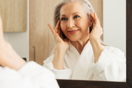 Close up of an aged woman with grey hair massaging her face in front of a mirror