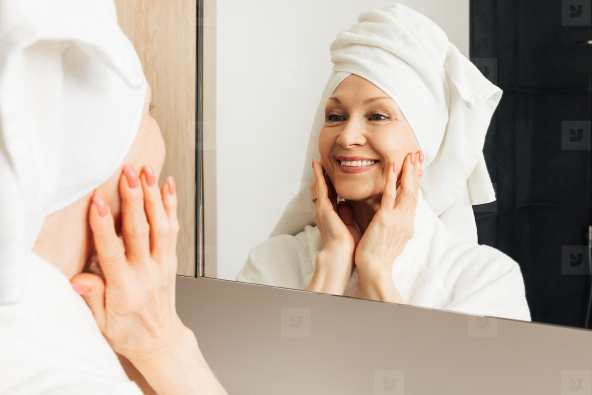 Portrait of a senior female with a towel on her head  Aged woman touching face in front on a mirror