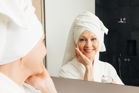 Portrait of a smiling senior woman applying facial treatment  Cheerful aged female with facial cream in bathroom