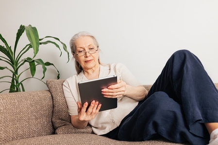 Aged female in eyeglasses lying on sofa reading from a digital tablet
