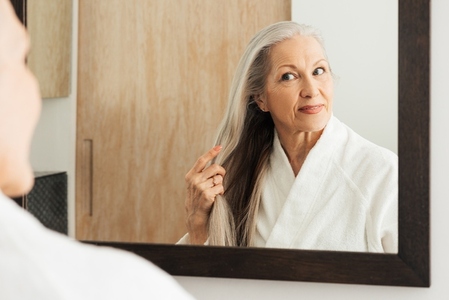 Senior woman examine her long grey hair while standing in front of a bathroom mirror