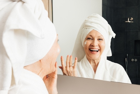 Laughing aged female with a wrapped towel on her head standing in front of a mirror in the bathroom