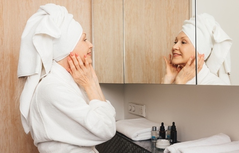 Side view of an aged woman with towel on her head massaging her face while looking at a mirror in the bathroom