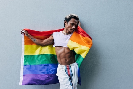 Stylish muscular guy leaning gray wall holds a rainbow LGBT flag