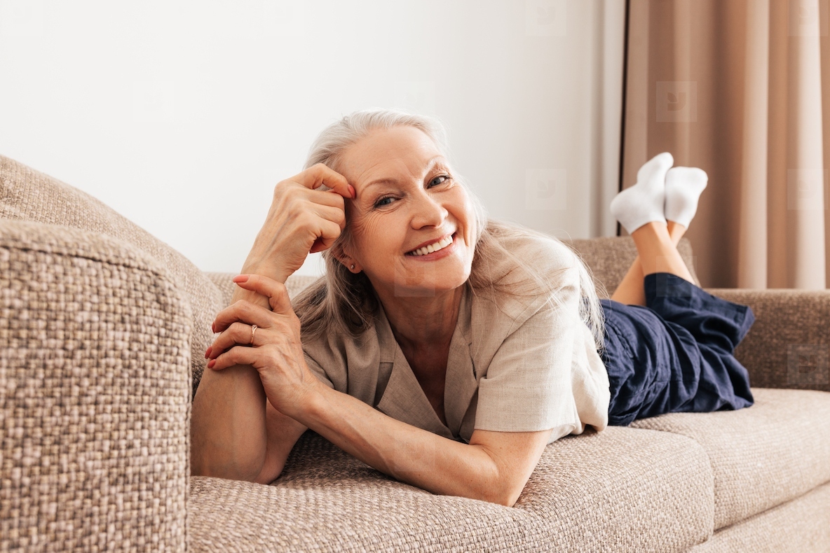 Portrait of a smiling aged woman with grey hair lying on a couch in living room looking at camera