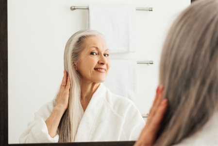 Senior woman touching her long grey hair and looking at a mirror in bathroom