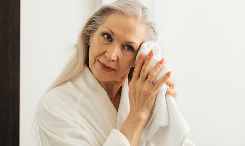 Aged woman with grey hair wiping her face in bathroom