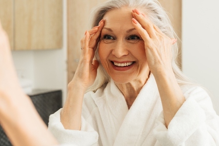 Close up of a happy aged woman touching her face with hands and looking at mirror