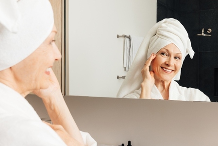Aged woman with wrapped towel on her head touching cheek looking at a mirror