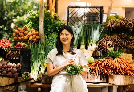 Cheerful Asian woman with a shopping bag standing at a farmers market  Young female standing at a stall in an outdoor market and looking at camera