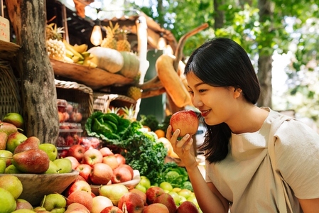 Asian woman smelling an apple at the counter on the outdoor market  Smiling female choosing fruit at a farmers market