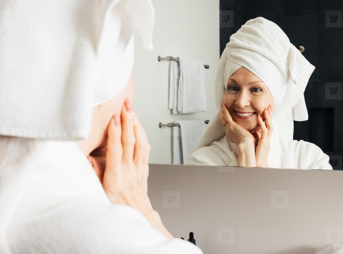 Cheerful woman admires her reflection in the bathroom mirror. Mature female touching her face with hands after the home spa