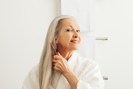 Senior adult woman combing her hair with a wood comb  Smiling female in a bathrobe taking care of her long gray hair