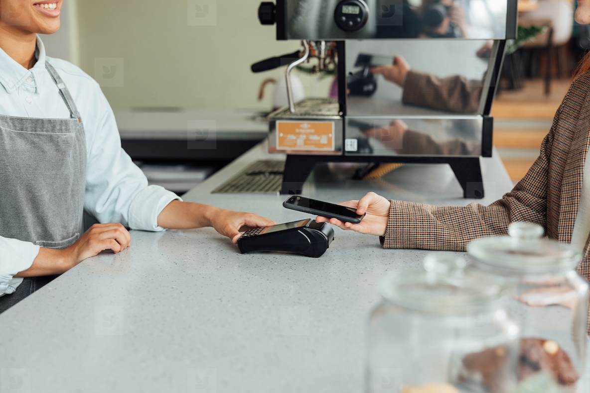Side view of a barista at counter receiving payment from a customer in a cafe. Unrecognizable female paying by mobile phone using NFC at the counter