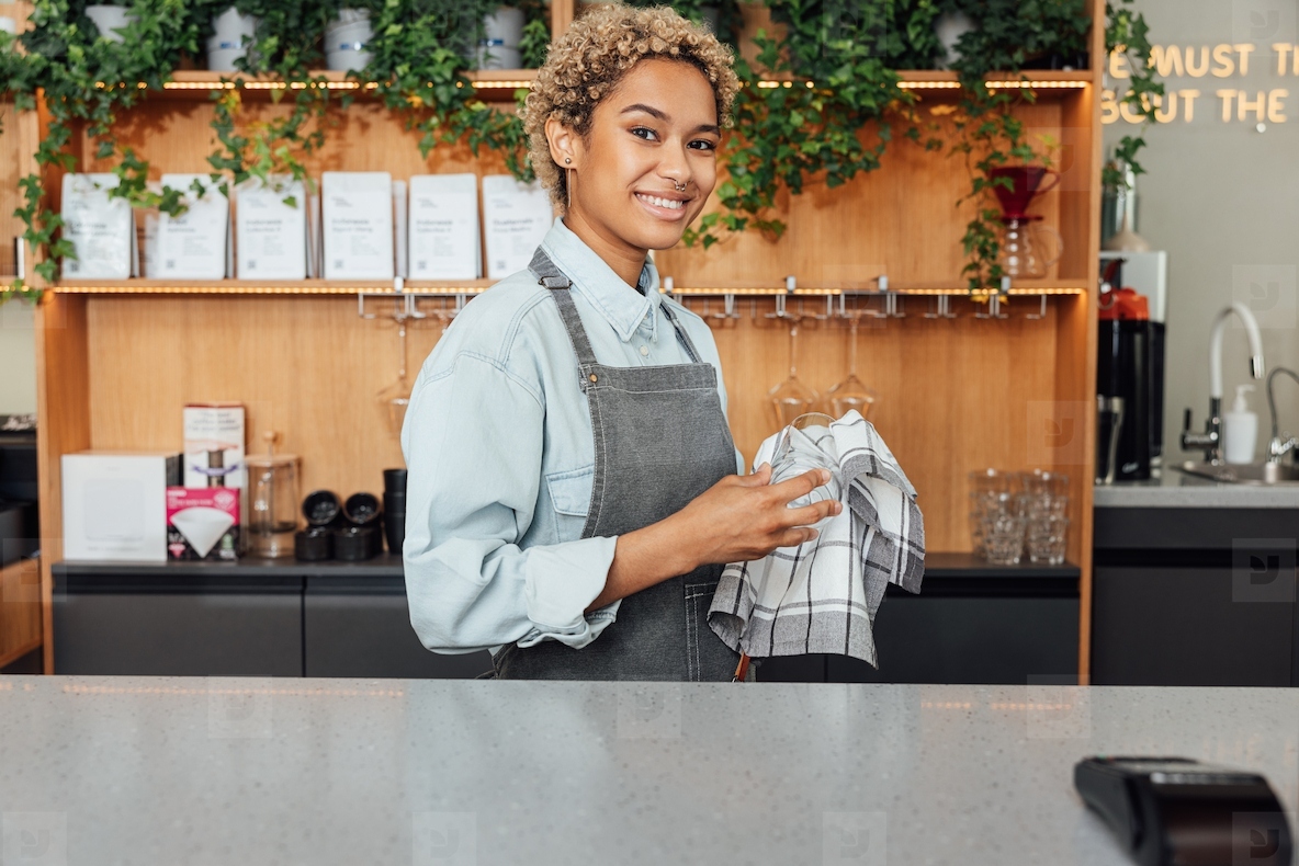 Smiling female bartender wiping glass with towel. Woman barista at counter in coffee shop