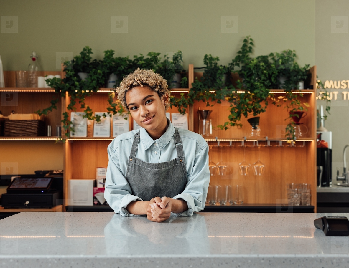 Barista in an apron with short curly hair leaning on a counter. Coffeeshop owner looking at camera