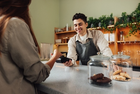 Smiling male barista holds a card machine while the customer pays by NFC  Barista in an apron receiving payment in a cafe at the counter