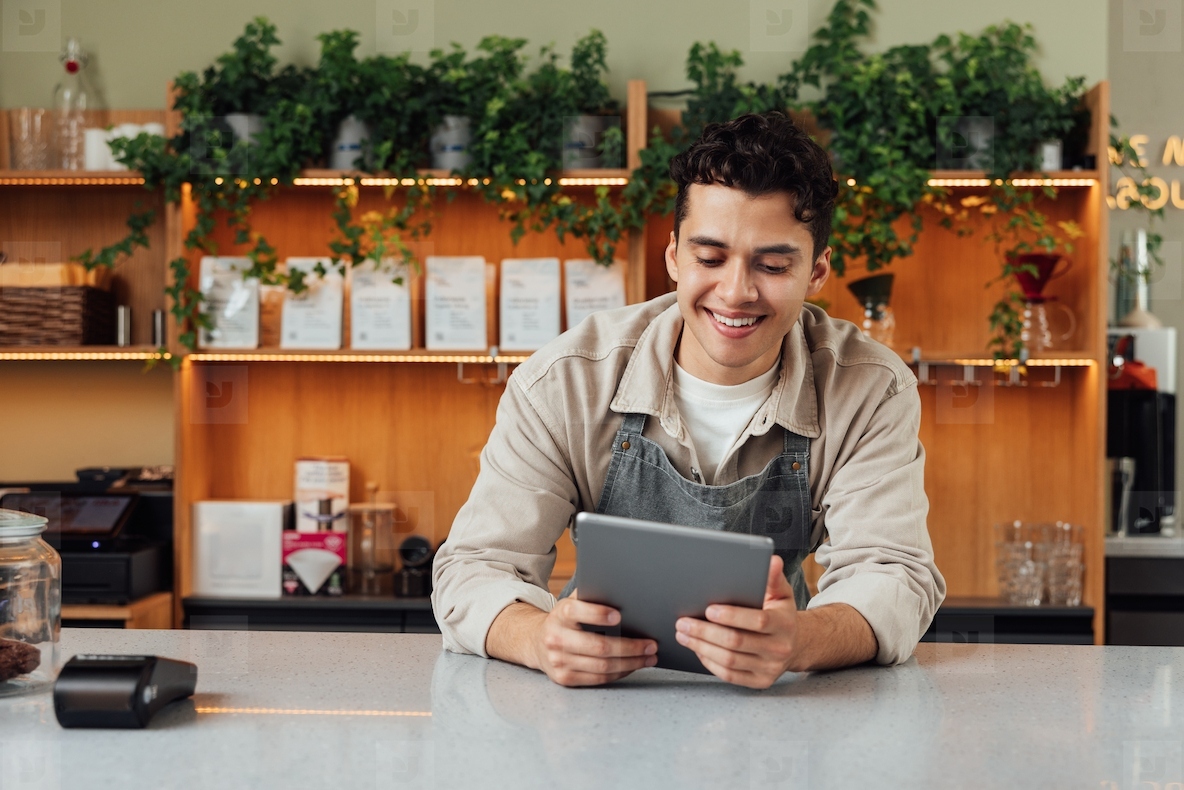 Smiling coffee shop owner at the counter with digital tablet. Male bartender in an apron leaning counter looking at a portable computer