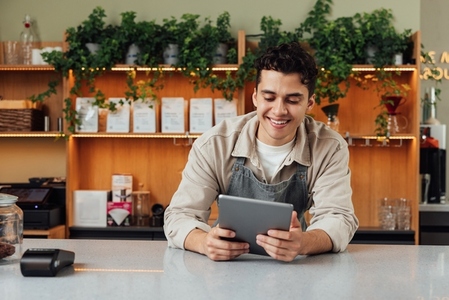 Smiling coffee shop owner at the counter with digital tablet  Male bartender in an apron leaning counter looking at a portable computer