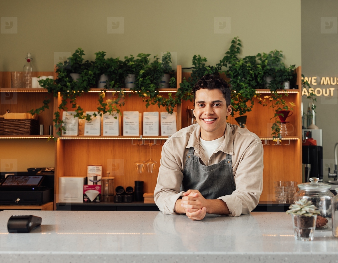 Smiling coffee shop owner in apron leaning a counter  Middle eastern bartender looking at camera