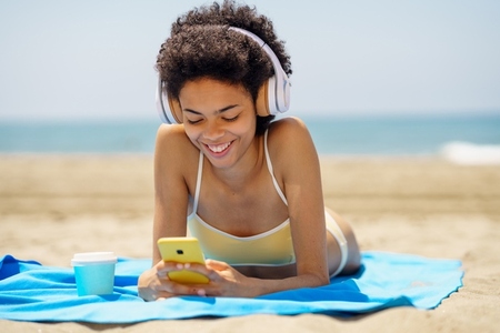 Delighted ethnic female tourist in headphones browsing smartphone on seashore