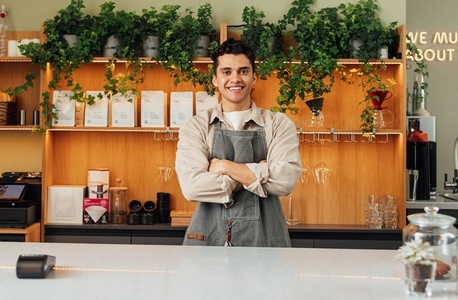 Confident male barista in an apron at the counter  Bartender with crossed hands looking at the camera and smiling