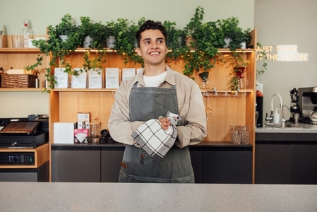 Smiling waiter in apron wiping hands with a towel while standing at the counter  Male barista in an apron with a towel