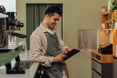 Side view of a smiling barista in an apron typing on a digital tablet leaning counter in a coffee shop