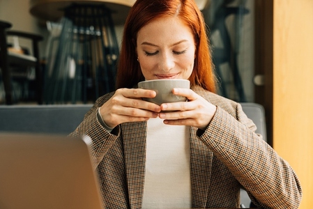 Close up of a young businesswoman holding a cup and smiling  Female with ginger hair enjoying her coffee in the morning in a cafe