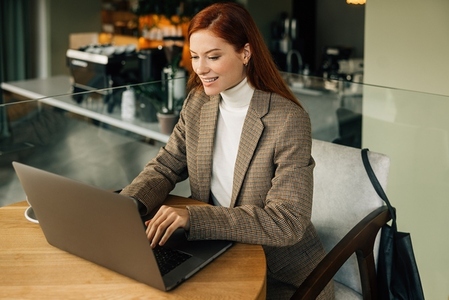 Smiling woman entrepreneur with ginger hair wearing blazer typing on laptop in the cafe