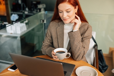 Businesswoman holding a cup of coffee and looking at laptop while sitting in cafe
