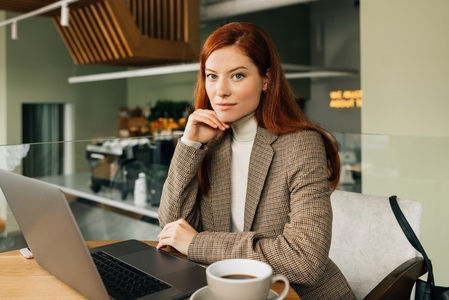 Portrait of a young businesswoman with ginger hair  Stylish female sitting at a table in a cafe with a laptop and coffee