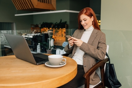 Smiling businesswoman with a smartphone in cafe  Female with ginger hair in formal wear sitting at a table