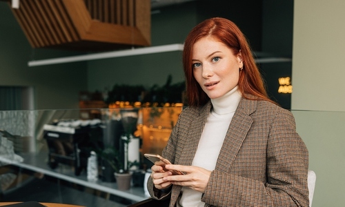 Portrait of a young businesswoman with ginger hair in a cafe  Female in formal wear holding a smartphone looking at camera