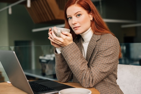 Portrait of a beautiful businesswoman with ginger hair holding a cup and looking at the camera while sitting in a cafe