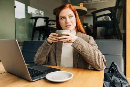 Woman with ginger hair holding a cup and looking at the window at a cafe  Businesswoman enjoying her coffee