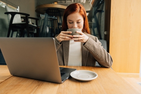 Smiling woman with ginger hair enjoying her morning coffee  Businesswoman holding a cup with closed eyes while sitting at a table